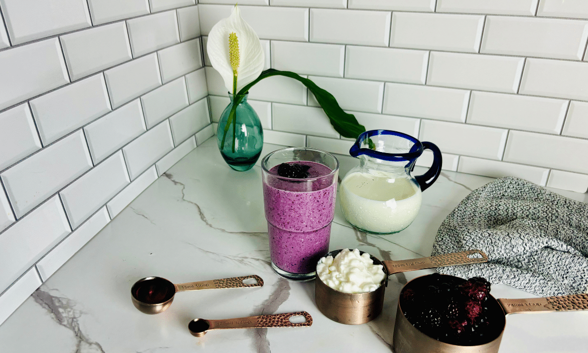 A purple berry cottage cheese smoothie topped with blackberries, next to a pitcher of milk, a cup of cottage cheese, frozen berries, and measuring spoons with maple syrup and vanilla.